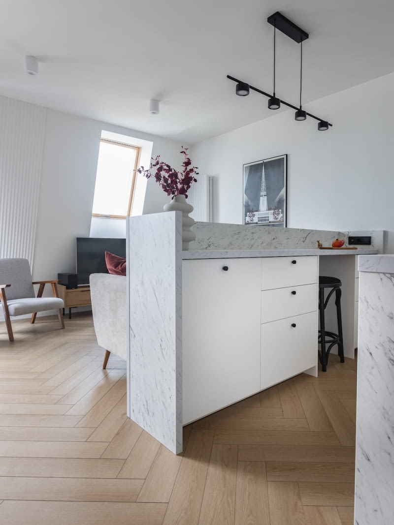 Jednym z założeń funkcjonalnych było zaprojektowanie przy aneksie kuchennym półwyspu. Miał on pełnić funkcję blatu roboczego i jednocześnie miejsca, przy którym można przysiąść i coś przekąsić. Nie bez powodu naprzeciwko znajduje się telewizor, na który również można zerkać, pracując przy blacie. Dodatkowo „wyciągnięcie” ścian półwyspu ponad poziom blatu optycznie oddziela kuchnię od salonu i skrywa nieład, który pojawia się podczas gotowania.