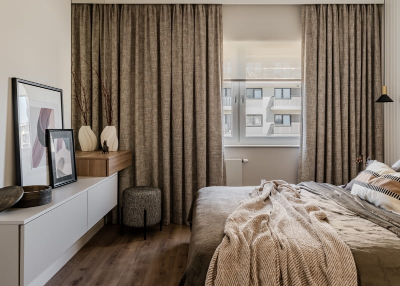 W sypialni okno zasłaniają brązowe, zaciemniające zasłony, ciemniejsze niż w pozostałych pokojach, utrzymane w tej samej tonacji co odcień paneli podłogowych.