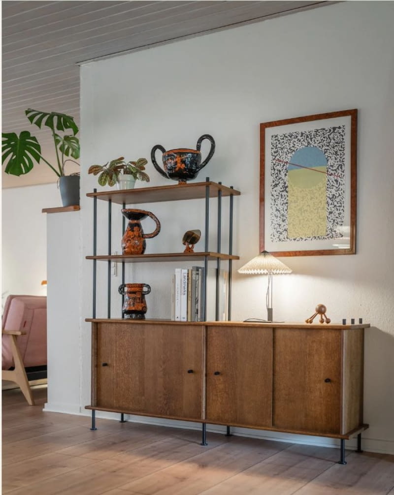 Asymetryczny układ regału Bava 160 daje idealny pretekst do uzupełnienia jego niższej części estetyczną dekoracją, tak by stworzyć przeciwwagę dla pionowej bryły mebla. Rama zawieszonego tu plakatu nawiązuje do drewna dębu, z którego powstał regał producenta TAMO.