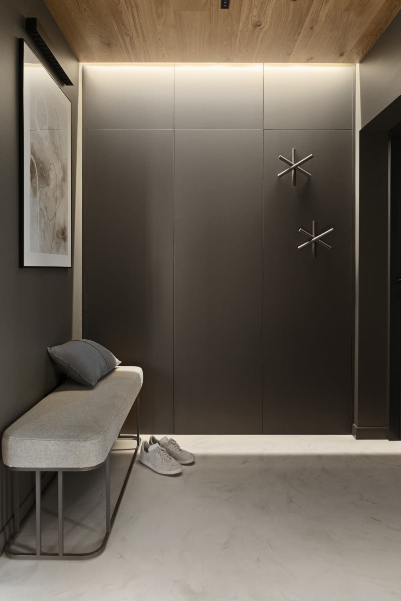 Bezspoinowa podłoga z żywicy w jasnym, neutralnym kolorze łączy wszystkie pomieszczenia parteru i pięknie podbija wszelkie barwy materiałów wykończeniowych. Zwłaszcza drewna czy grafitowych płyt meblowych Forner Velvet Ultra Matt, z których wykonano fronty szaf na ubrania w wiatrołapie i jego ściany widziane od strony salonu, a także zabudowę meblową w kuchni. Na tle ciemnej powierzchni idealnie prezentuje się zrobiona na zamówienie ławka ustawiona w wiatrłoapie. Podstawa siedziska ma formę nawiązującą do stolika kawowego w salonie, a wybrany materiał tapicerski to ten sam, którego użyto na salonowej kanapie. Wykonane na zamówienie z tych samych materiałów elementy wyposażenia tworzą spójną całość.