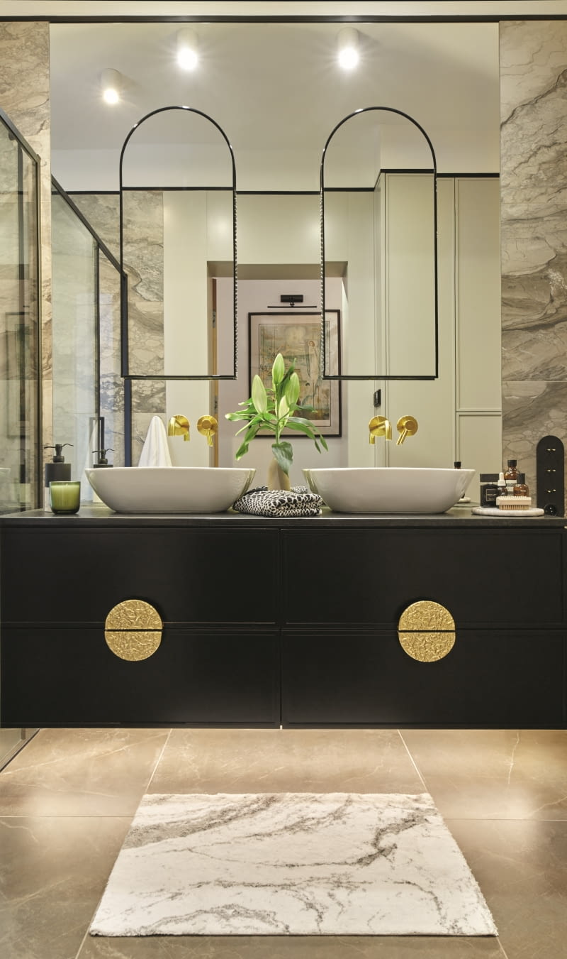 Centralnym punktem długiej łazienki rodziców jest wspaniała, czarna, z czarnym, kamiennym blatem szafka umywalkowa i lustrzana aranżacja wokoło niej. Wyjątkowym detalem są mosiężne uchwyty meblowe od Pap Deco, które nadają charakteru zabudowie. Równie oryginalnym pomysłem pozostają zamocowane na tafli lustra dwa mniejsze, łukowe lustra, które są przyporządkowane do każdej z dwóch umywalek. Odbija się w nich zabudowa meblowa wokół toalety.