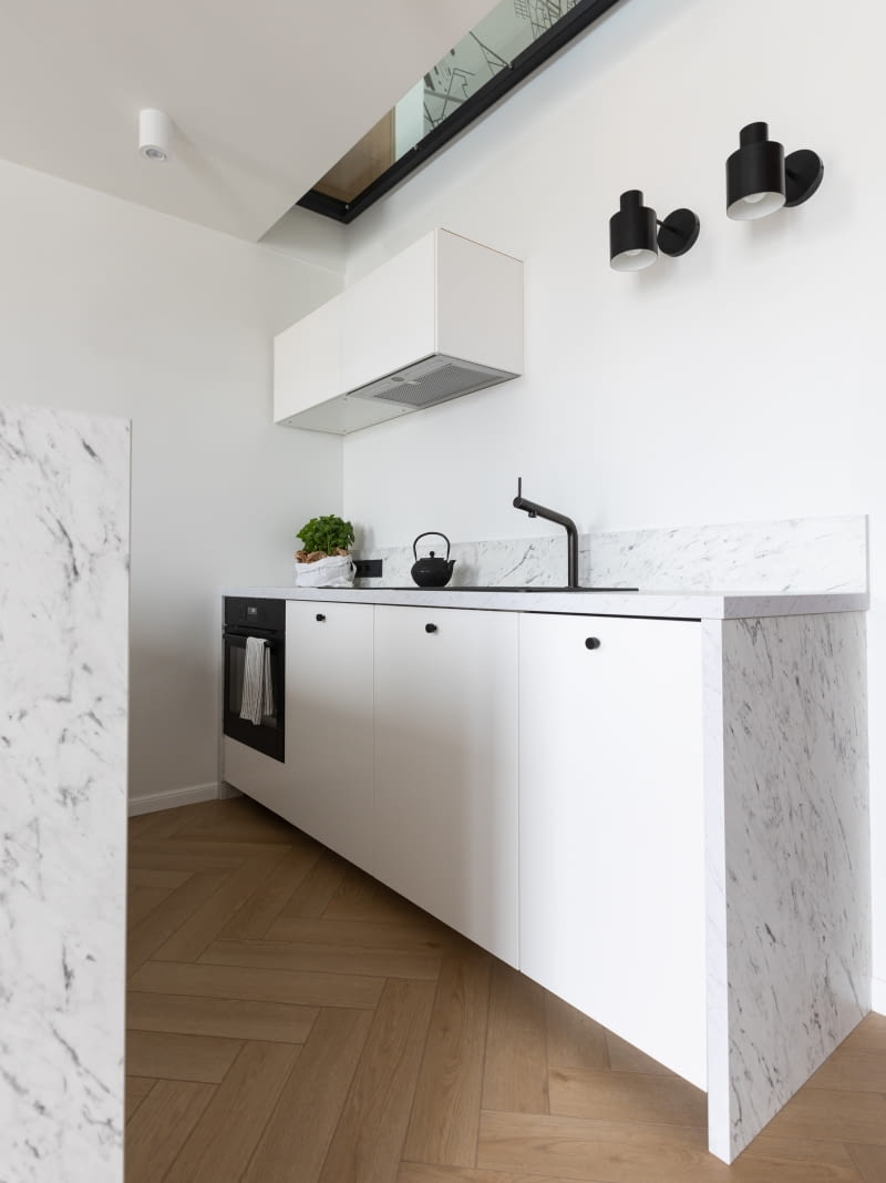 Meble kuchenne pochodzą ze sklepu Ikea, seria Metod. Dla projektantki ważne było, że w kolekcji znajdują się szafki w dwóch głębokościach. Część kuchni przy ścianie powstała ze skrzyń 60 cm, a półwysep z płytszych – 40 cm. Półwysep zajmuje więc mniej cennego miejsca. Blaty oraz obudowę szafek wykonano z płyt laminowanych Egger, imitujących marmur Carrara. Lampa wisząca nad blatem półwyspu – Nordlux, czarne kinkiety – Maxlight Black, gałki meblowe – Skandynawskie Uchwyty.