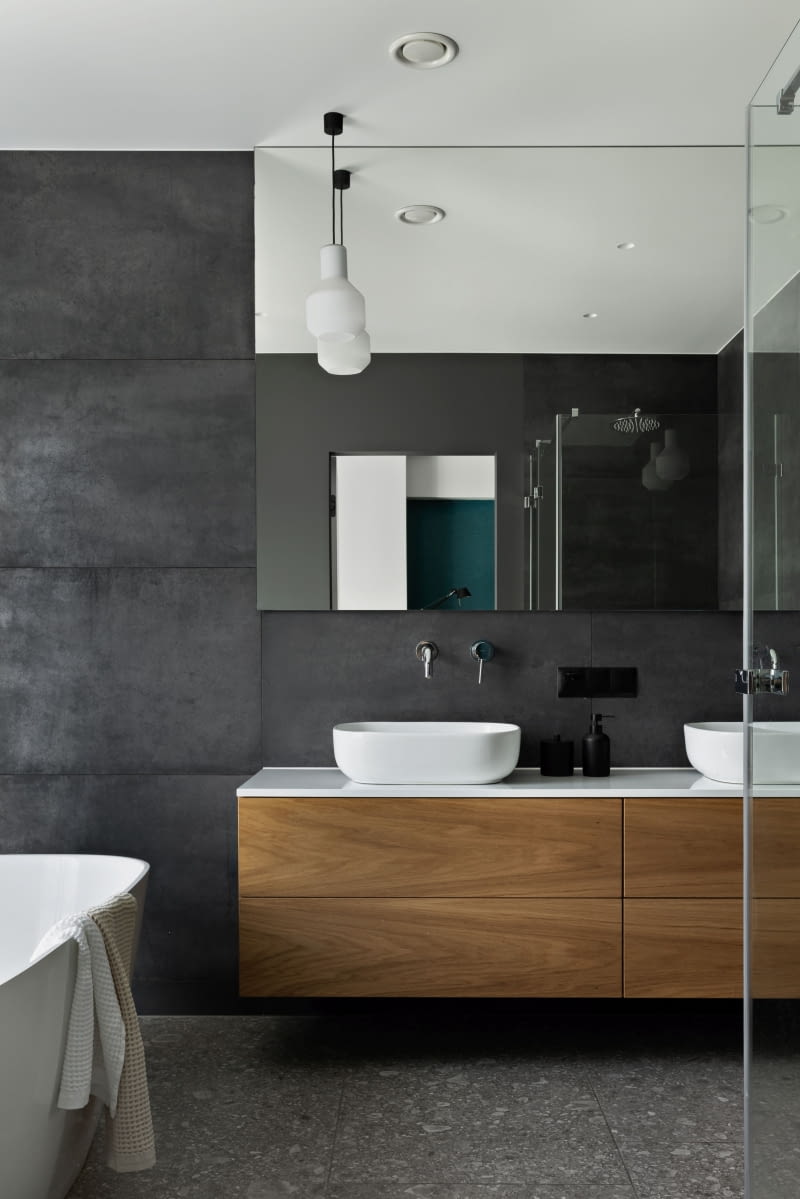 W łazienkach również zadbano o spójność z aranżacją całego domu. W łazience rodziców antracytowe płytki Mirage zestawiono z dębowym fornirem – tym samym, który pojawia się w kuchni. Na podłodze, podobnie jak w pozostałych pomieszczeniach, ułożono gres Ergon ze wzorem kamienia Ceppo di Gré. Właścicielom zależało na jednolitej posadzce scalającej wszystkie strefy domu. Choć początkowo rozważali drewno, ostatecznie wybrali gres – praktyczny, łatwy w utrzymaniu i doskonale współpracujący z ogrzewaniem podłogowym. W łazienkach również zadbano o spójność z aranżacją całego domu. W łazience rodziców antracytowe płytki Mirage zestawiono z dębowym fornirem – tym samym, który pojawia się w kuchni. Na podłodze, podobnie jak w pozostałych pomieszczeniach, ułożono gres Ergon ze wzorem kamienia Ceppo di Gré. Właścicielom zależało na jednolitej posadzce scalającej wszystkie strefy domu. Choć początkowo rozważali drewno, ostatecznie wybrali gres – praktyczny, łatwy w utrzymaniu i doskonale współpracujący z ogrzewaniem podłogowym.