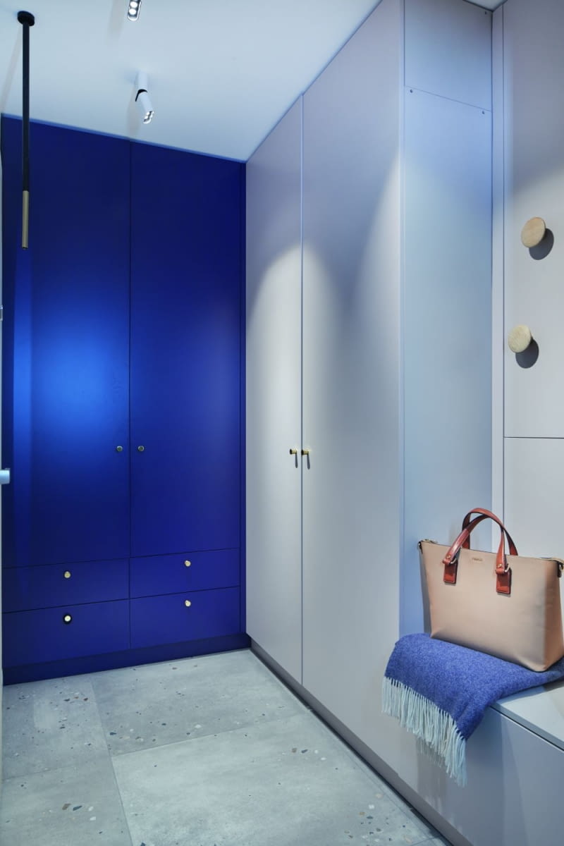 Przy wejściu do mieszkania zaprojektowano zabudowę meblową wykończoną w tych samych kolorach, co w innych pomieszczeniach. Pierwotnie w miejscu niebieskiej szafy było WC, ale gospodarze zdecydowali, że wolą mieć tu podręczną garderobę. Powstały więc dwie pokaźne szafy. Znalazło się też miejsce na tapicerowane siedzisko i dekoracyjne, duże uchwyty-wieszaki. Podłogę zdobią te same co w kuchni płytki 80 x 80 cm Marmara.