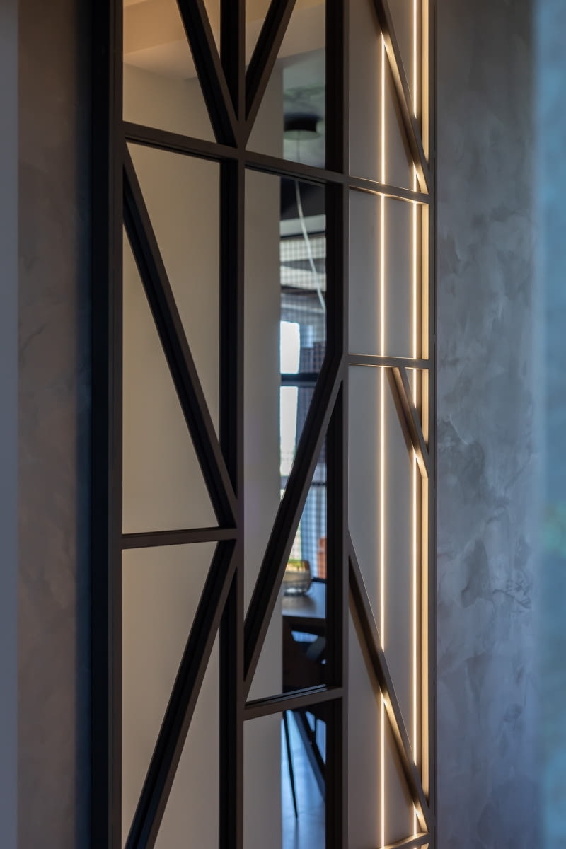 Monotonię korytarza łączącego część dzienną z nocną urozmaica ozdobny panel. Architekta zaprojektowała go ze stalowych, malowanych na czarno profili i lustra. Panel podświetlony listwami LED’owymi, zwłaszcza wieczorem, przy rezygnacji z oświetlenia górnego, dyskretnie rozjaśnia ciemny hol, budując przyjemny nastrój.