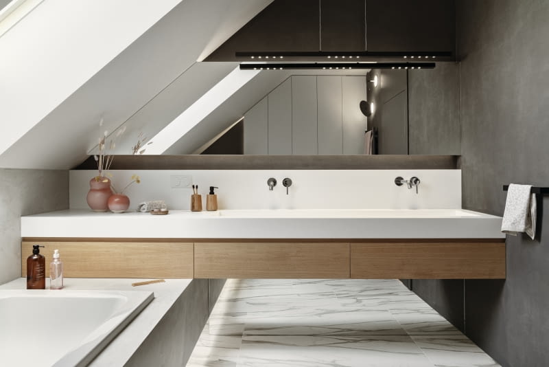 W aranżacji ogólnodostępnej łazienki na piętrze, z której korzystają głównie dzieci, uwagę przyciąga ciekawie zaprojektowana szafka umywalkowa. Jej podwieszana, rozpięta między ścianami bryła przylega do prostopadle ustawionej wanny w obudowie, jakby się na niej opierając. Szafka dźwiga blat zintegrowany z długą, rynnową umywalką, wykonaną z białego Corianu. Nad umywalką zamontowano dwie ścienne baterie.