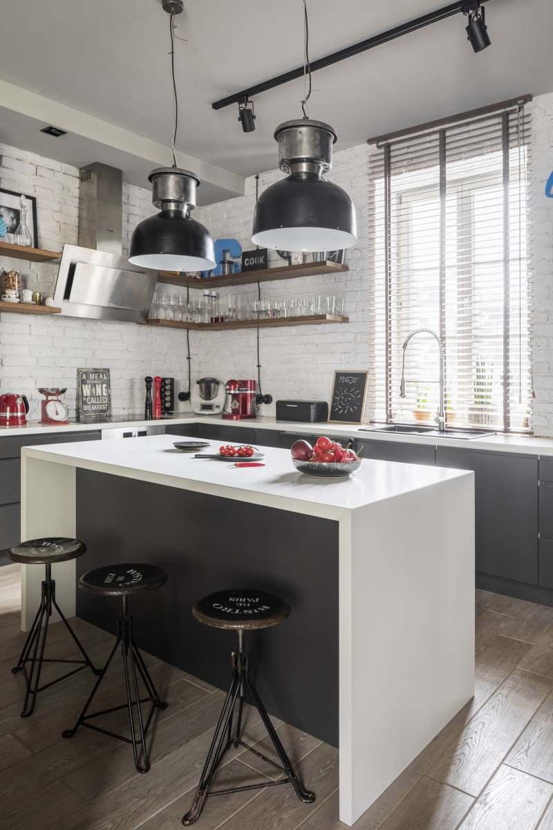 Zintegrowaną z częścią dzienną kuchnię wyposażono w niskie szafki oraz wyspę kuchenną, unikając dominowania strefy gotowania w otwartej przestrzeni. Lodówkę umieszczono w wysokiej zabudowie oddzielającej kuchnię od strefy wejścia. Nowoczesne materiały wykończeniowe: lakierowane płyty MDF w kolorze grafitowym oraz blaty z jasnego spieku kwarcowego urozmaicają industrialną aranżację. Dzięki nim loftowe wnętrze nie jest przeciążone przyjętą stylistyką.