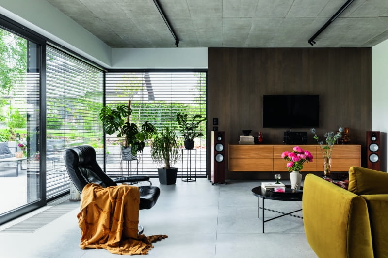 Beton wkracza śmiało również do salonu, bo z niego wykonano sufit, choć klimat tej części parteru kształtują głównie duże aluminiowe przeszklenia, bardzo szczelne, a przede wszystkim wpuszczające do środka duże ilości naturalnego światła – no i ogrodowe widok