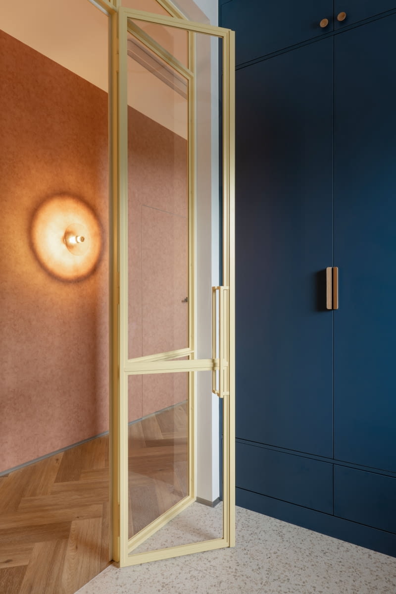 Przedsionek oddziela od korytarza szklana ściana osadzona wraz z drzwiami w stalowych, żółtych profilach – kolor ten przewija się w całym domu. Ściany korytarza i wybranych pomieszczeń pokryto tapetą Caselio z kolekcji Beton 2, o delikatnej, betonowej strukturze, zastosowaną w różnych odcieniach, tak by zachować spójność wzoru i jednocześnie zróżnicować przestrzeń poprzez barwy. Do korytarza wybrano wersję w ceglastym tonie.