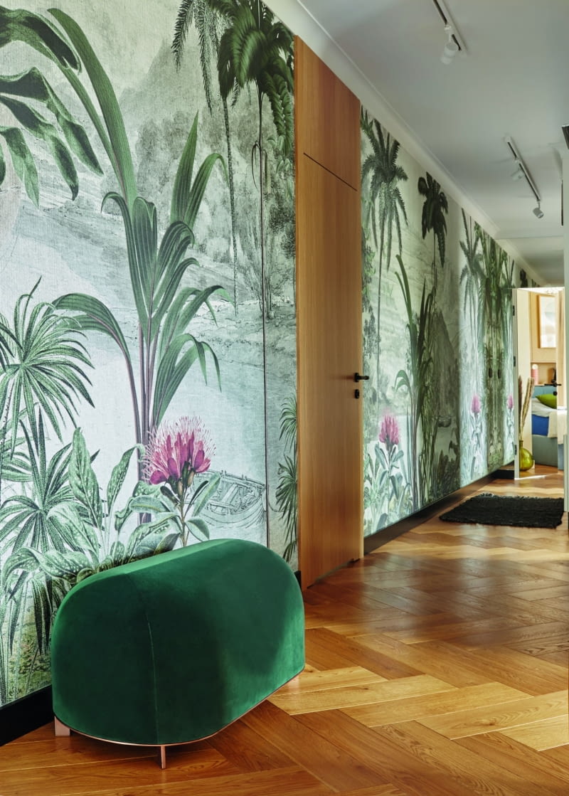 Długa ściana korytarza ciągnącego się przez całe mieszkanie była zdaniem projektantki od początku idealnym miejscem na tapetę. Przyklasnęli temu domownicy, gdyż bardzo lubią ten rodzaj wykończenia. Magda chciała podkreślić długość holu intensywnym wzorem. Razem z panią domu wybrała tapetowe inspiracje, a firma dekorami.pl stworzyła wzór idealnie dopasowany do wyobrażeń obu pań.  Na samym końcu korytarza umieszczono lustrzane drzwi do sypialni rodziców, co podkreśla jeszcze bardziej jego długość. Intensywna tapeta, lustrzane oraz fornirowane drzwi nadają korytarzowi dekoracyjny rys. Pracuje na to również dopracowany w szczegółach zielony puf Nurt/Noti Studio na subtelnych nóżkach z drewna.