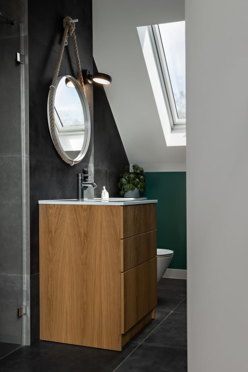 W łazience na piętrze na jednej ze ścian i na podłodze ułożono antracytowe płytki o wyglądzie betonu, a we wnęce prysznicowej o kilka tonów jaśniejsze – grafitowe. Szafkę umywalkową stolarze wykończyli dębowym fornirem, tym samym, którego użyto w pozostałych meblach w domu. W łazience na piętrze na jednej ze ścian i na podłodze ułożono antracytowe płytki o wyglądzie betonu, a we wnęce prysznicowej o kilka tonów jaśniejsze – grafitowe. Szafkę umywalkową stolarze wykończyli dębowym fornirem, tym samym, którego użyto w pozostałych meblach w domu.
