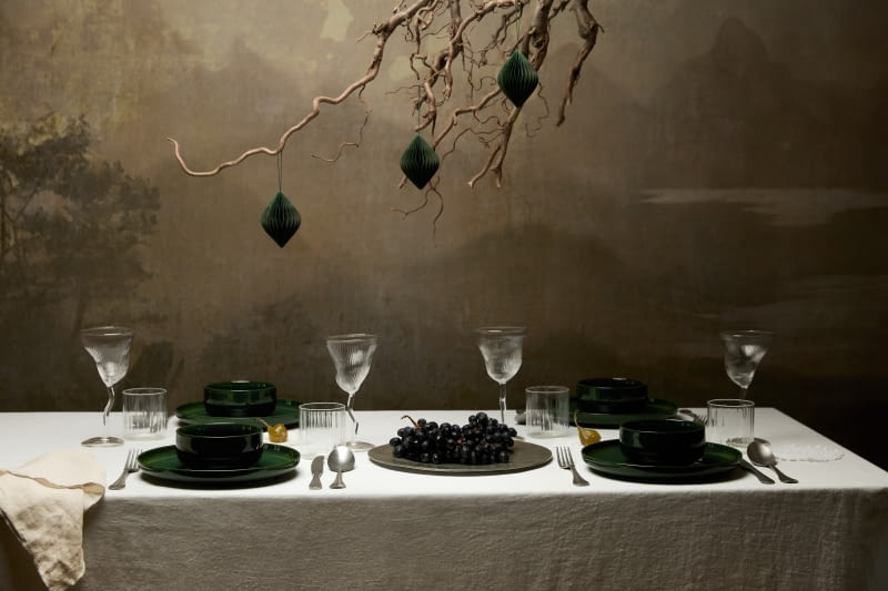 Prostą aranżację stołu wzbogacono podwieszoną nad nim podłaniczką o bardzo nowoczesnej formie. Zamiast zielonej wykorzystano nagą gałązkę, na której zawisły trzy identyczne bombki.