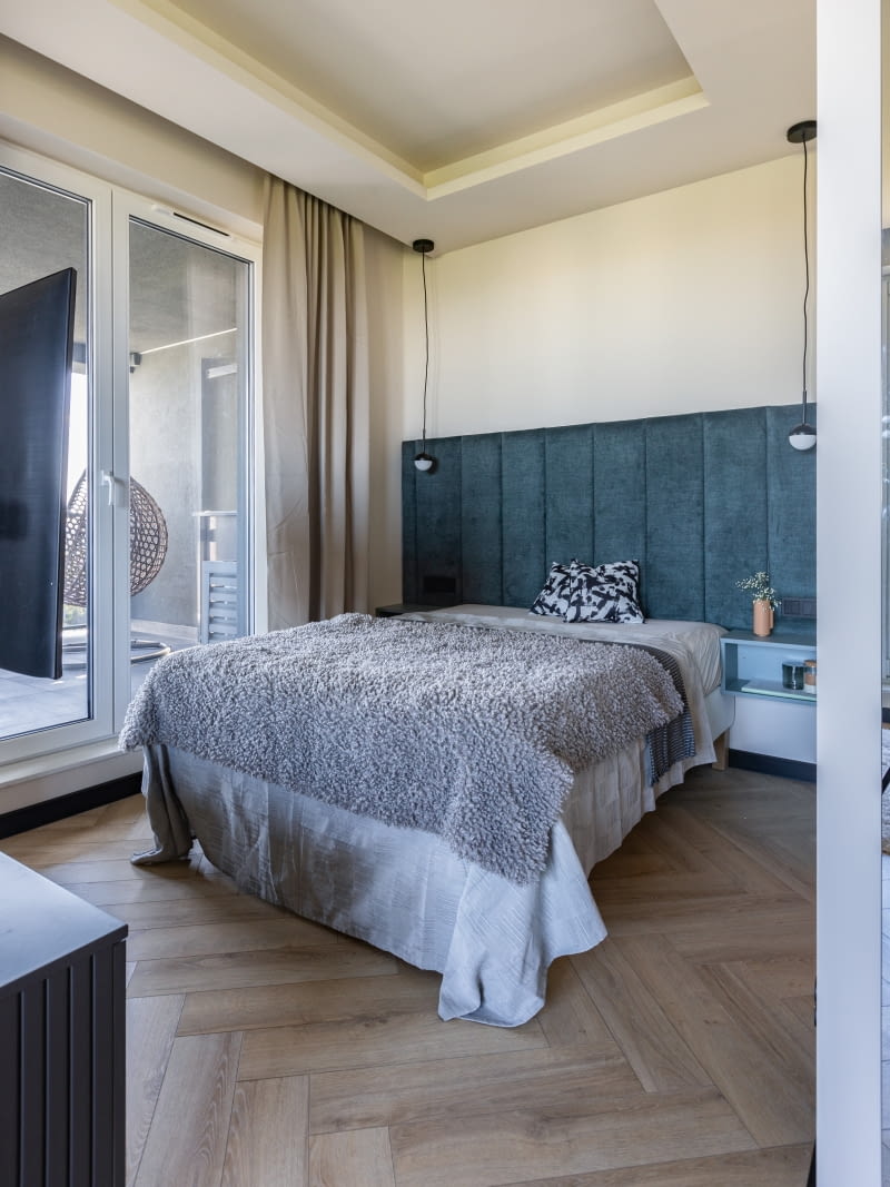 Najważniejsze w sypialni jest oczywiście łóżko, podkreślone efektownym, tapicerowanym zagłówkiem w kolorze burzowego morza. Nie mniej ważne, decydujące o wygodzie wyposażenie, są umieszczone po bokach materaca zgrabne, otwarte półki do odłożenia książki, telefonu, czy kubka. Wiszące przy łóżku lampy do czytania mają formę czarno-białych kul.