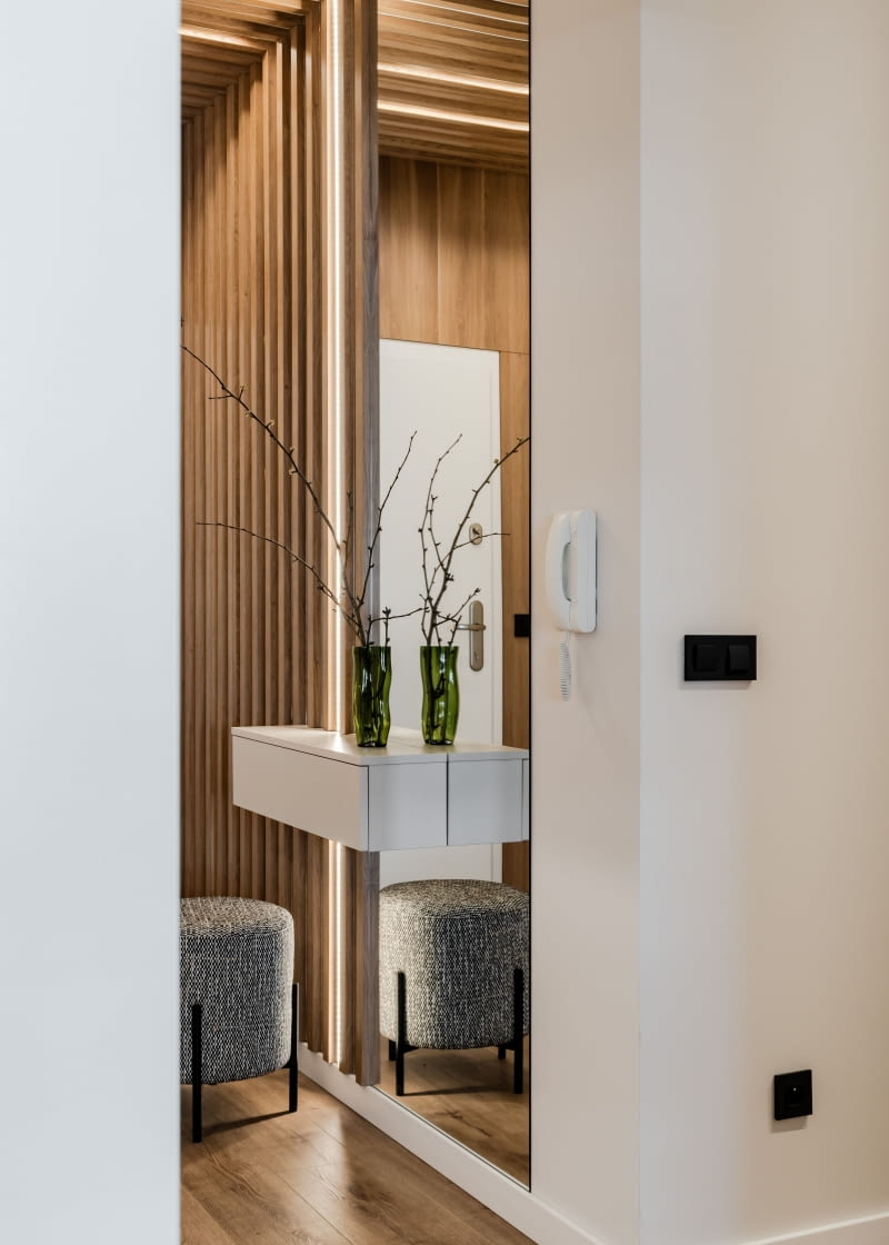 W korytarzu pojawiają się lamele wykonane przez stolarza z płyty Egger Orzech Carini Naturalny – tej samej, co w łazience. Pomiędzy lamelami zastosowano oświetlenie LED. Zaprojektowano miejsce na odłożenie kluczy oraz lustro.