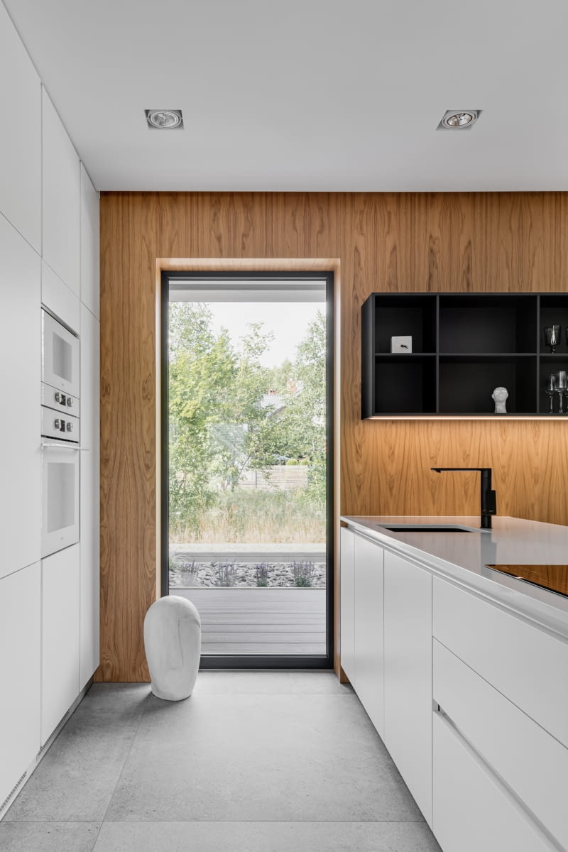 Ściana wykończona drewnem stanowi interesującą ramę dla widoku za oknem i przełamuje białą zabudowe meblową.