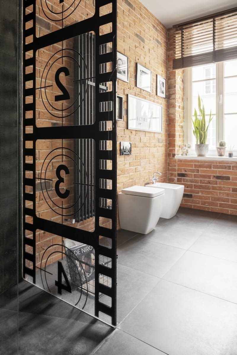 W łazience, tak jak w całym mieszkaniu, zachowano zastaną, nietynkowaną cegłę ceramiczną. Udało się ją oczyścić i uzupełnić brakujące spoiny zaprawą nawiązującą do tej oryginalnej. Surowa, naturalnie niedoskonała powierzchnia kontrastuje z nowoczesną, betonową ścianą pokrytą płytami zamówionymi u producenta klinikabetonu.pl oraz z gładką, podłogą, wykończoną płytami gresowymi w szarym, lekko melanżowym kolorze.