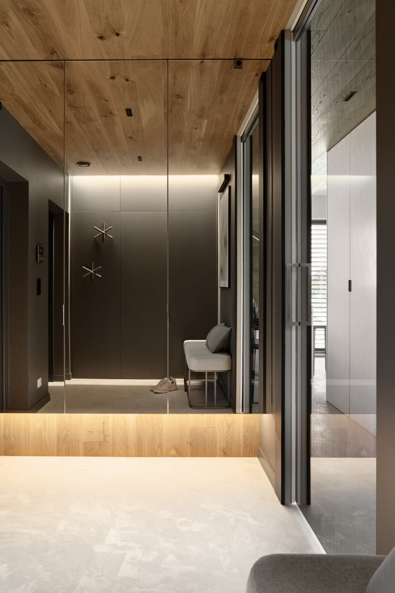 W wiatrołapie za ścianą z luster i odbijających się w nich z naprzeciwka grafitowych frontach zabudowy meblowej przewidziano miejsce na kurtki i buty. Z pomieszczenia przechodzi się do salonu przez drzwi przesuwne chowane w ścianie. Ponieważ są przeszklone i wysokie na 2,5 m, zapewniają w niewielkim przedsionku poczucie przestrzeni oraz wgląd w strefę salonu i ogrodu.