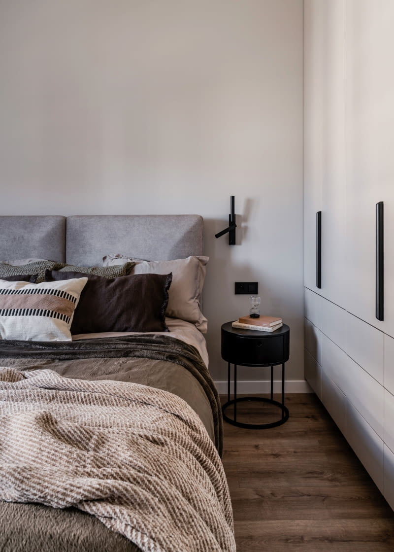 Wygodne, pasujące wizualnie do sypialni łóżko Senpo Hamilton dobrano w beżowoszarej tapicerce. Towarzyszą mu po bokach dwa czarne stoliki z szufladami oraz lampy, z jednej strony wygodny podczas czytania czarny kinkiet kierunkowy,  a z drugiej model wiszący.