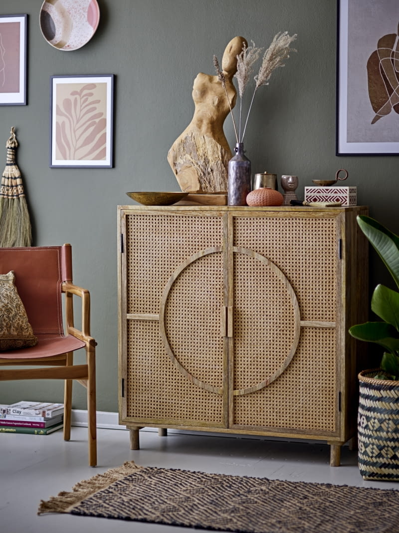 Dekoracje ustawione na komodzie tworzą zróżnicowaną kompozycję – od wysokiej rzeźby, przez średni wazon z trawami, po niskie pudełko czy miseczkę. Ciepłe, ziemiste odcienie wyraźnie odcinają się od ciemnej ściany, budując przytulny nastrój.