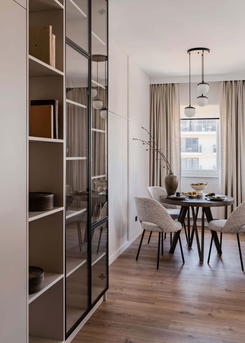 Widok z korytarza w stronę części dziennej kończy starannie dopracowana kompozycja okrągłego stołu jadalnianego ustawionego na tle okna. Spokojną linię długiego przejścia przełamuje zaprojektowana tu witryna na książki i zastawę stołową. Dzięki niej korytarz nabrał dynamiki i wyrazistego charakteru.