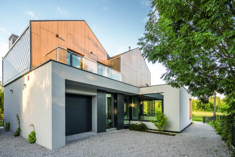 Strefę wejścia do budynku (na prawo od wjazdu do garażu) bardzo skutecznie ukryto przed wzrokiem osób postronnych – usytuowano ją  w sporym wycięciu w bryle, osłaniającym tę część domu od drogi dojazdowej, biegnącej w dole działki