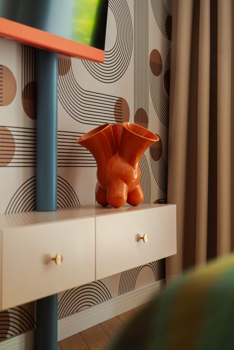 Metalowe rury malowane na niebiesko zasłaniają mocowania telewizora, ale stanowią też jeden z elementów kompozycji z wykorzystaniem geometrycznych kształtów. Prowadzone od sufitu do podłogi przecinają bryłę poziomo zawieszonej, niewysokiej, różowej szafki. Na blacie pomarańczowy wazon z oferty Comforty.