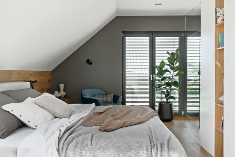 W sypialni rodziców nie mogło zabraknąć obecnego w całym domu drewna. Na podłodze oraz na ścianie za łóżkiem architekci zastosowali deskę warstwową dębową, taką samą jak na sufitach na parterze. Ciemny materiał na blacie szafek nocnych to szczotkowany marmur Pietra Grey w kolorze grafitowym, który wykorzystano do obudowy kominka i na blacie stolika kawowego w salonie.