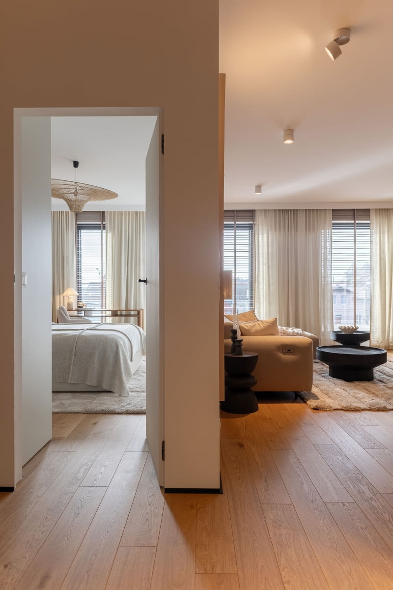 Strefę wejścia, w pierwotnym projekcie dewelopera zamkniętą ścianami i drzwiami, otwarto na część dzienną, dzięki czemu już od progu widać jasny salon z wysokimi oknami. O ich oprawę zadbała Natalia z salonbellacasa.pl – identyczne drewniane żaluzje i beżowe zasłony zastosowane w każdym oknie nadają wnętrzu spójny styl. W salonie pomiędzy oknami, pod zasłonami, ukryto grzejnik  oraz jednostkę klimatyzacji, tak by nie przyciągały uwagi i nie zaburzały odbioru całej koncepcji.