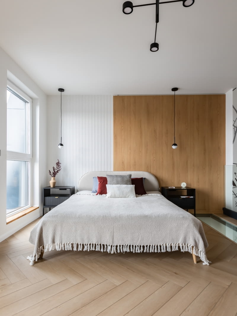 Ścianę przy łóżku wyłożono dębową okładziną i sąsiadującym z nią białym panelem ryflowanym 3D Orac Decor, który pojawia się w również w innych pomieszczeniach. Pomysłowe płaszczyzny stanowią ciekawe tło dla łóżka w beżowej tapicerce marki Optisofa oraz dla czarnych szafek nocnych z Menito, również wzbogaconych motywem ryflowania na frontach szuflad. Lampy na suficie i wiszące przy łóżku – Nordlux.