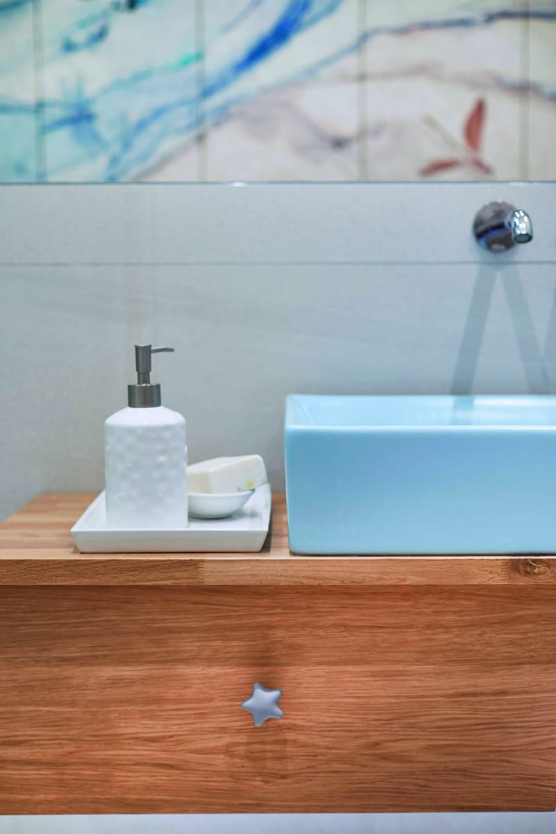 W łazience dzieci projektantka dobrała stonowane barwy, ale nawiązujące niebieskim kolorem do aranżacji salonu. Łagodny wystrój ożywiają płytki nad wanną oraz naklejona na szafki tapeta od Wonderwall ze wzorem ważek. Obydwa rozwiązania przedstawiono w niebieskich odcieniach, przywodzących na myśl wodę i fale. Niebieska jest też umywalka i uchwyty szuflad w szafce.