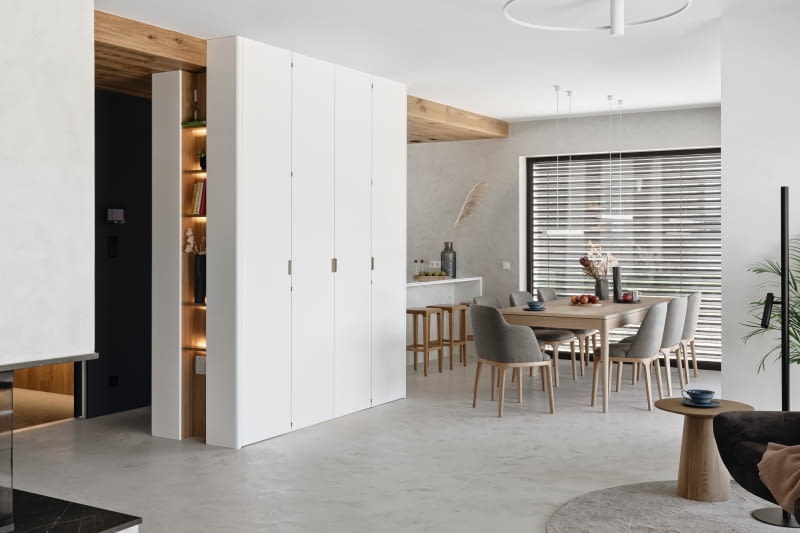 Kuchnię dyskretnie cofniętą względem części dziennej zasłania zabudowa meblowa wykonana wokół istniejącego tu fragmentu ściany konstrukcyjnej. Po otwarciu białych, o pełnej wysokości frontów widać eleganckie, podświetlone wnętrze wykończone fornirem dębowym. To idealne, poręczne miejsce obok stołu na zastawę stołową. Dębowy stół marki Selfia kupiono w sklepie 9design.pl, a krzesła w IWC Home.