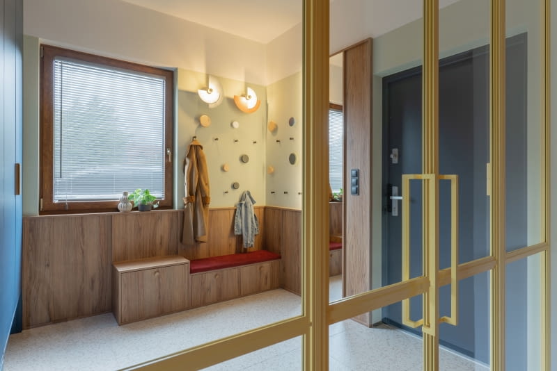 Zawieszone nad siedziskiem na różnych wysokościach haczyki ułatwiają dzieciom odwieszanie kurtek. Ułożono je w kompozycję z drewnianymi, okrągłymi dekoracjami ściennymi oraz dwoma kinkietami hiszpańskiej marki Aromas del Campo.