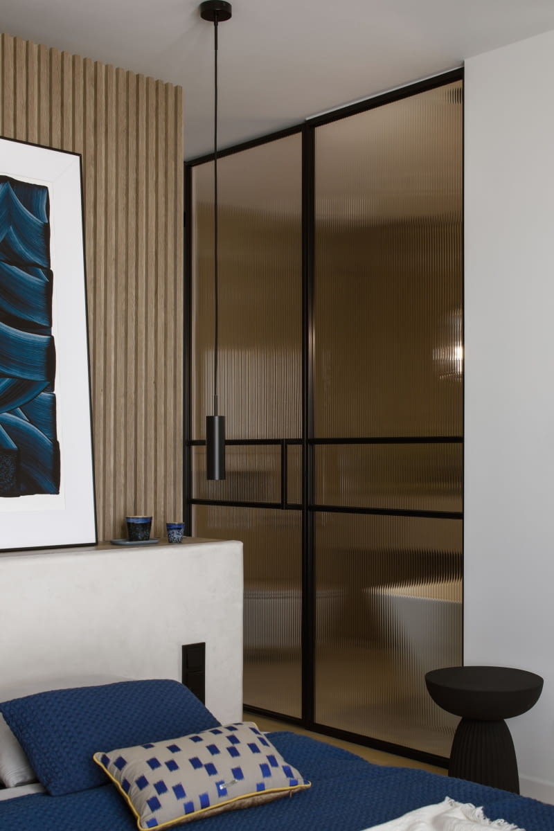 Wysokie do sufitu drzwi w czarnych, metalowych ramach, obejmujących tafle szkła typu flutes, powtórzono przy przejściu z salonu do sypialni rodziców,  a także do ich osobistej łazienki. Dzięki lekko transparentnej przegrodzie pomieszczenia przenikają się, tworząc wrażenie większej, nieograniczonej przestrzeni.