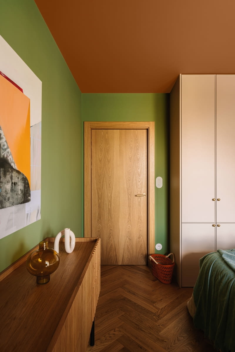 W sypialni dominuje spokojna, relaksująca zieleń, skontrastowana z ciepłym, rudym sufitem, który otula przestrzeń. Całość dopełnia naturalne drewno obecne na parkiecie, meblach wykonanych przez stolarza i drzwiach marki Entra.
