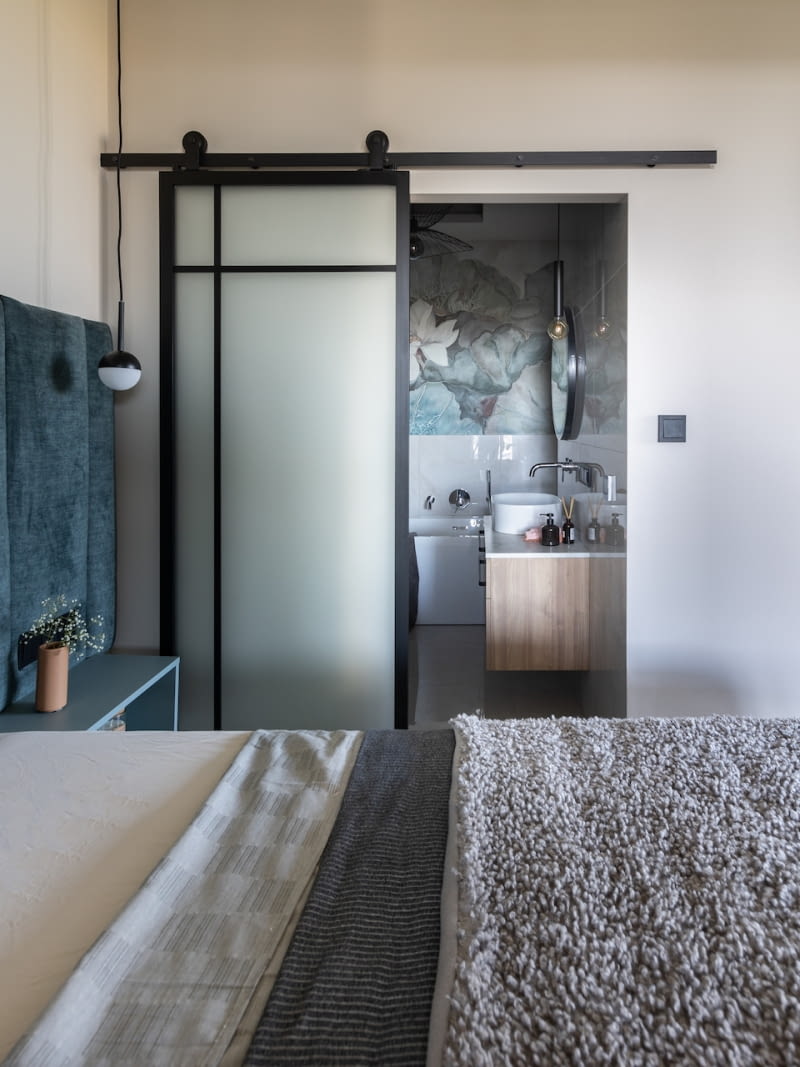 Przesuwne drzwi w metalowej ramie prowadzą do łazienki domowników. Wypełnione mlecznym szkłem sprawiają wrażenie wizualnie lekkich i doświetlają cennym światłem łazienkę. Jednocześnie są nieprzezierne, co daje mieszkańcom poczucie prywatności. Ponadto przesuwny model eliminuje problem z tradycyjnym otwieraniem skrzydła, które w tym miejscu musiałoby się opierać o łóżko. Sama stylistyka drzwi podkreśla związek z wystrojem dziennej części mieszkania – surowej, inspirowanej bliską sercu Rafała estetyką loftową.