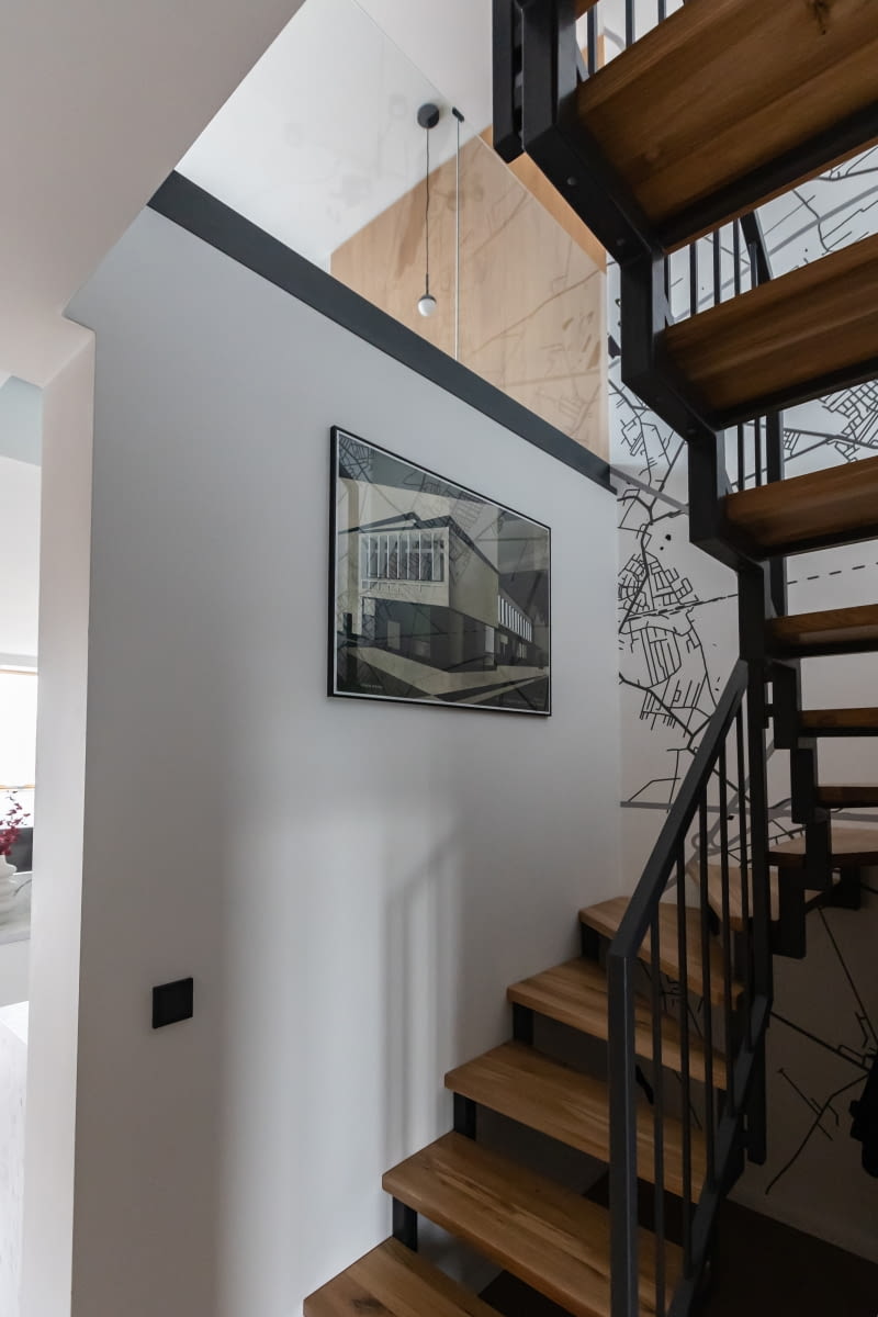 Ażurowe, niezabudowujące przestrzeni schody prowadzące na piętro zamówiono u producenta Mr. Steel. Stopnie wykonano z jesionu zabezpieczonego bejcą, a konstrukcję z malowanej w antracycie stali. Biała ściana klatki schodowej eksponuje plakat Ryszarda Kai, przedstawiający poznańską Galerię Miejską Arsenał.