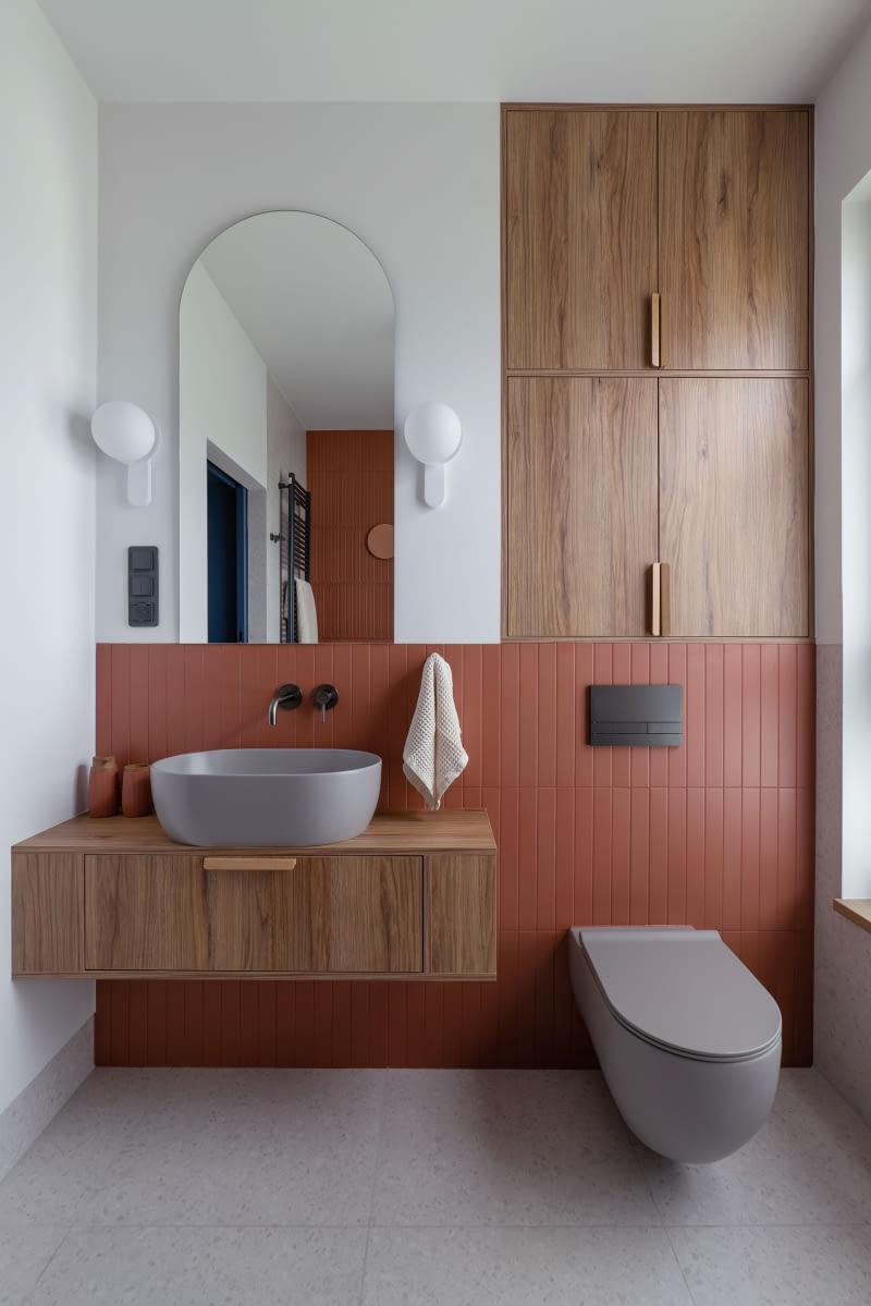 W łazience rodziców wykorzystano dwa rodzaje płytek – w kolorze ciepłej, lekkiej szarości od producenta Terrazzo oraz ceglaste od Equipe. Białe kinkiety  z motywem łuku dopasowanego do kształtu lustra to model od Ummo Lighting, a we wszystkich łazienkach zastosowano armaturę Omnires.