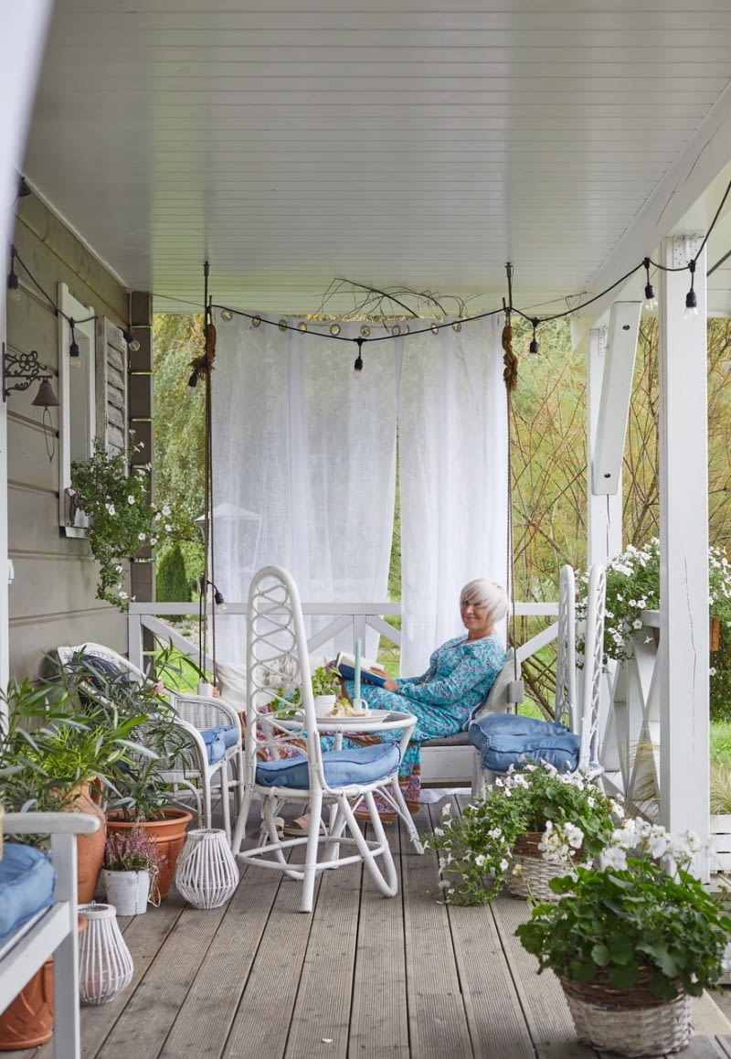 Wystrój werandy zapowiada klimat panujący we wnętrzu domu – swobodny, dający poczucie bezpieczeństwa i komfortu, bazujący na naturalnych materiałach i kolorach. Połączenie szarej elewacji i desek na podłodze werandy z bielą jej sufitu, barierki oraz drewnianych i rattanowych mebli wprowadza nastrój jak z prowansalskich posiadłości, skąpanych w słońcu i sezonowej zieleni.