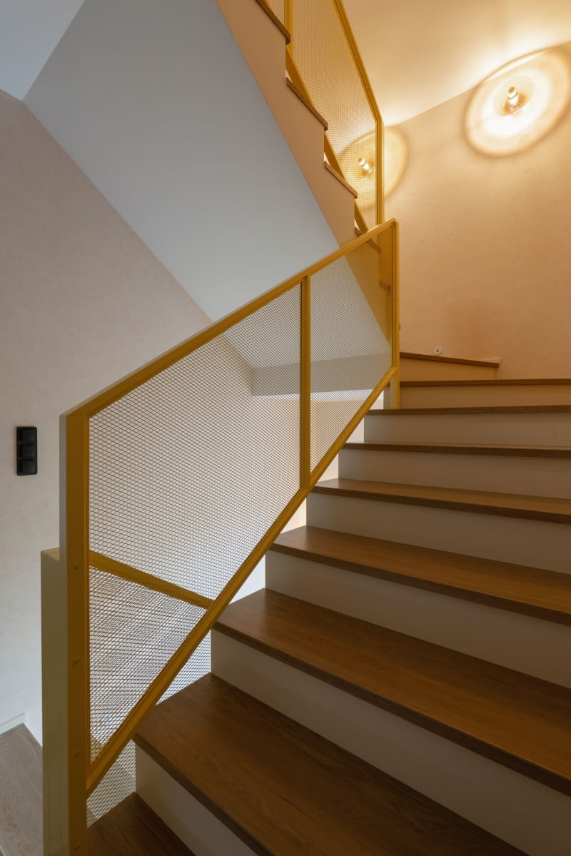 Betonowe schody wykończono naturalnym drewnem – stopnie są dębowe, a podstopnice również drewniane, ale malowane na odcień złamanej bieli. Na ścianach powtórzono tapetę o betonowej strukturze, tym razem w beżowym kolorze. Jej przecieraną fakturę podkreśla światło okrągłych kinkietów Aromas del Campo, takich samych jak w korytarzu na parterze. Całość ożywia żółta, metalowa barierka – nawiązanie do szklanych drzwi przedsionka.
