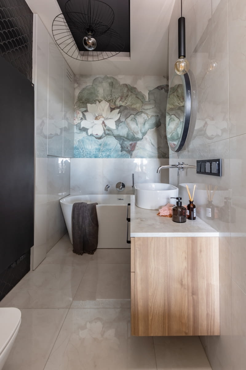 Kwiatowa fototapeta Wall & Deco w osobistej łazience gospodarzy przyciąga wzrok i czyni wnętrze eleganckim. Wrażenie to podkreśla także przyścienna wanna o owalnym kształcie naśladująca wolno stojące modele oraz opalizujące płytki Tubądzina na ścianach i podłodze, jak również blat szafki umywalkowej z konglomeratu Technistone. Jasne powierzchnie przełamuje czerń wnęk – sufitowej i ściennej. Tę drugą zdobi czarna mozaika Raw Decor. Na jej tle czarny, prosty grzejnik staje się niemal niewidoczny.