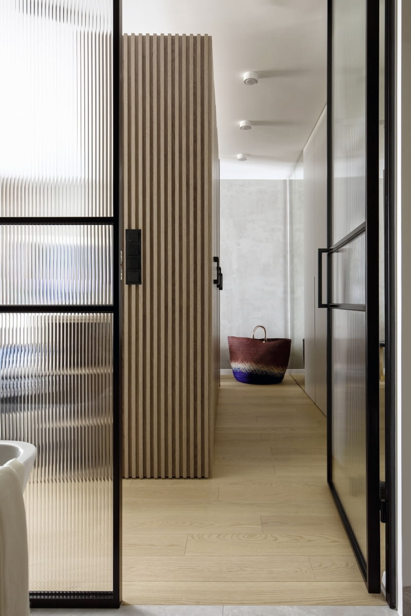 Pionowe frezowania ornamentowego szkła w drzwiach nawiązują do linii lamelowych paneli obudowujących szafę garderoby. Taka wertykalna struktura wizualnie podwyższa pomieszczenie. Ciekawostką jest, że bryła szafy nie sięga sufitu. Zabieg ten pozwala na większy dostęp naturalnego światła z sypialni  do garderoby.