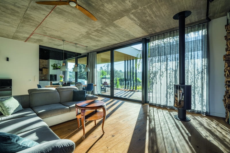 Połączone ze sobą salon, jadalnia i kuchnia pełne są akcentów przemysłowych, na które składają się zarówno betonowy sufit, uzyskany z szalunku  stropu, jak i stylowa żeliwna koza. Elementami ocieplającymi ów wizerunek są wygodne meble z miękkim, szarym obiciem i „antykwaryczny” stolik  kawowy, a gdzieś pośrodku tych klimatów lokuje się duma właścicieli – robiony na zamówienie regał na całą ścianę, wykonany z metalu i drewna