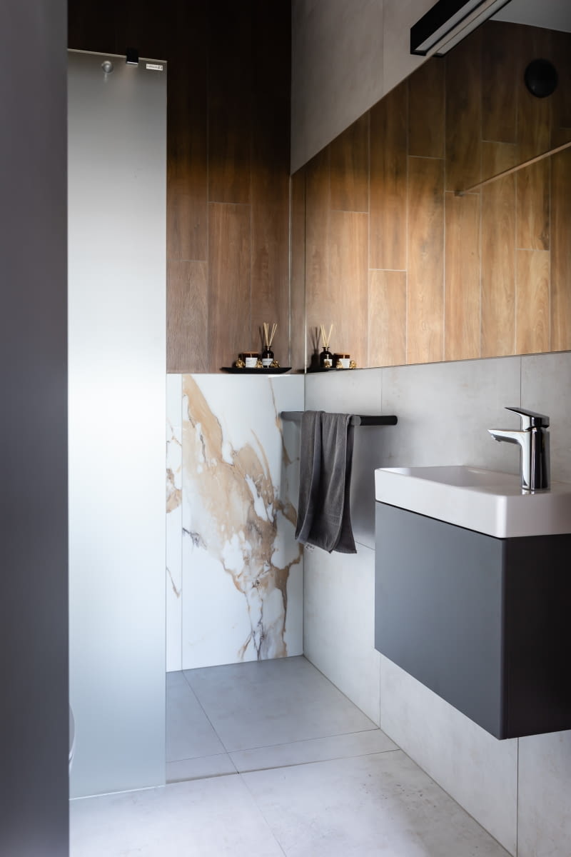 Wystrój łazienki gościnnej nawiązuje do strefy dziennej. Mają wspólną podłogę wykończoną tymi samymi, szarymi płytkami 60 x 60 cm marki Tubądzin. Pojawia się tu także ciemna szarość pod postacią szafki podumywalkowej i jednej ze ścian oraz czarne detale – uchwyt na ręcznik, oprawa lampy i osprzęt elektryczny. Płytki firmy Ceramica Limone imitujące drewno wraz z gresem Geotiles o wyrazistym wzorze kamienia wprowadzają do aranżacji szczyptę luksusu, który zachęca do relaksu.