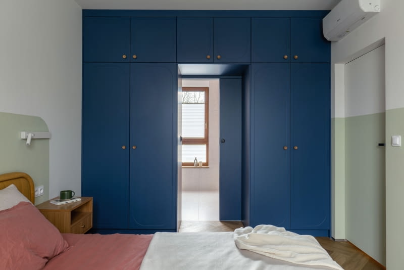 Przejście z sypialni rodziców do ich prywatnej łazienki ujęto w wysoką zabudowę ciemnoniebieskich szaf na ubrania. Umieszczone centralnie w jej bryle drzwi, pomalowane w kolorze frontów, zlewają się z ich rytmem i wyglądają jak kolejny moduł zabudowy. Skrzydło to model przesuwny, chowany w kasecie ukrytej w ścianie za szafą.