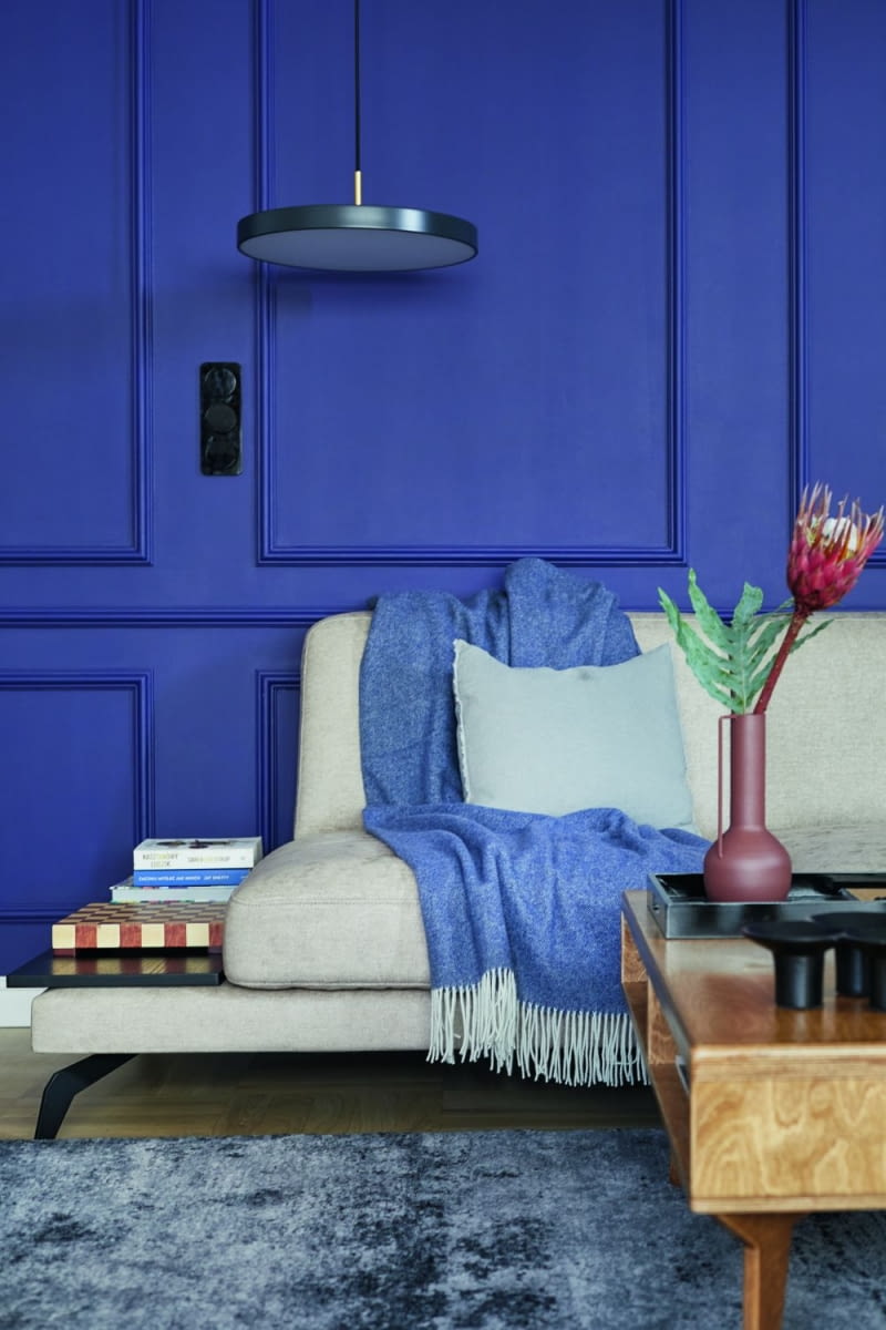 Niebieska ściana z ozdobną sztukaterią w salonie od początku była w planach Hanny i Łukasza. Wygląda elegancko i dodaje charakteru wnętrzu. Projektantka uwzględniła ten kolor również w innych pomieszczeniach, dzięki czemu aranżacja całego mieszkania jest spójna. Barwa świetnie komponuje się z kolorem drewna, czernią i szarością. Jest obecna również w dodatkach, na przykład tekstyliach o różnych odcieniach błękitu. Ułożone na sofie: pled, lniane i welurowe poduszki pochodzą z @yourest_studio.