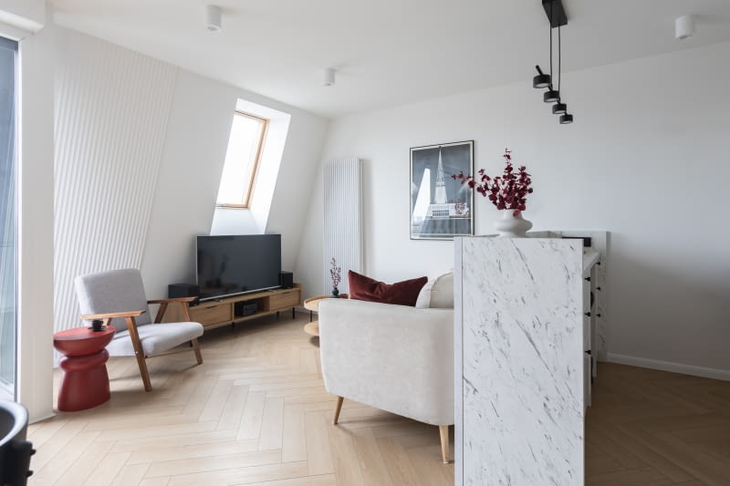 W aranżacji całego mieszkania chodziło o uzyskanie wrażenia jak największej przestrzeni. Stąd niewiele mebli wolno stojących, jasna kolorystyka, funkcja przechowywania skoncentrowana w kubikach. Nieliczne, najważniejsze dekoracje dotyczą głównie miasta Poznania – przy sofie plakat z iglicą targów poznańskich (autor – Ryszard Kaja). Sofa – marka Optisofa, szafka RTV i stolik burgundowy – Górna Półka, fotel – meble.pl