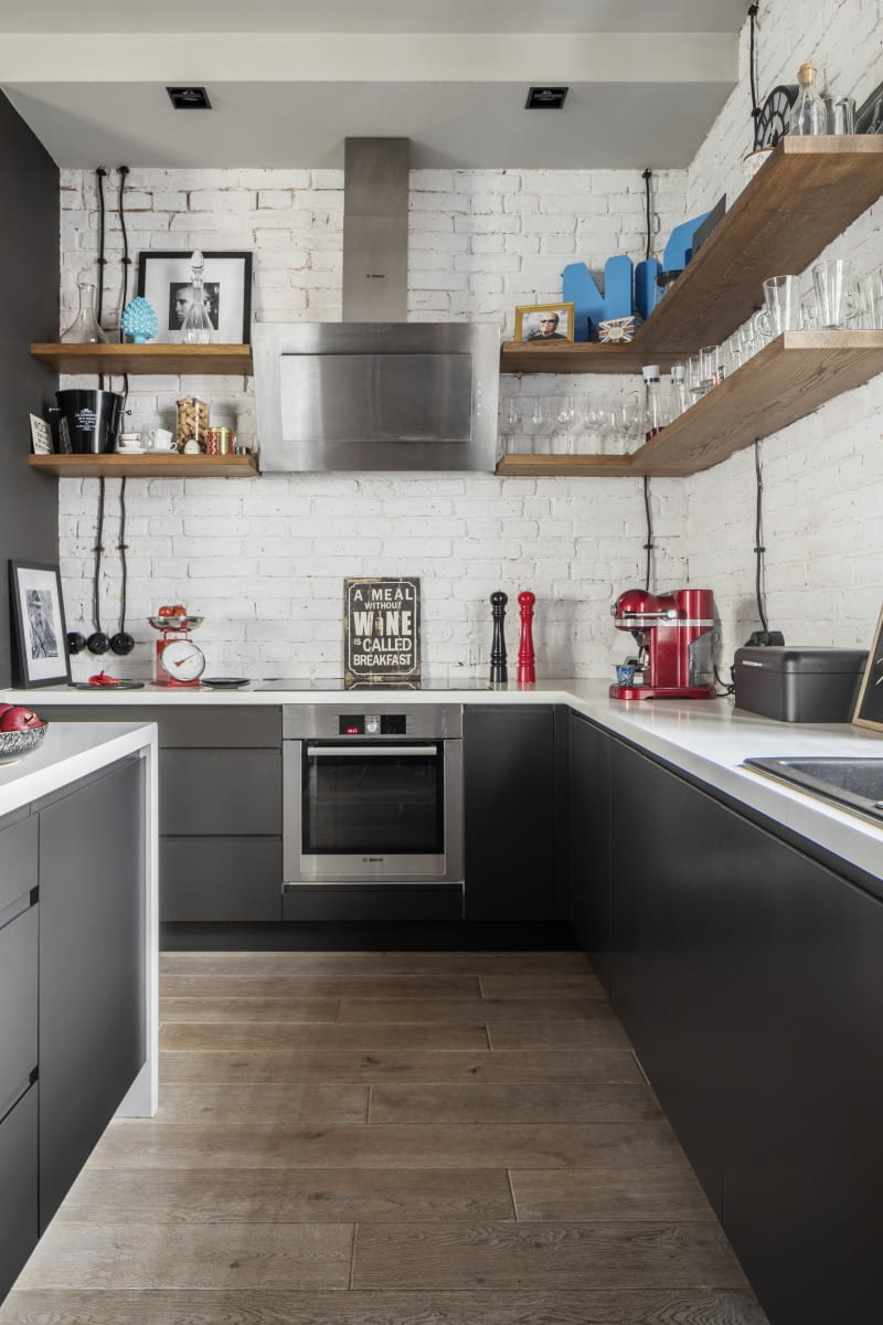 Wystrój kuchni harmonizuje ze strefą dzienną. Zapewnia to wykończenie podłogi w całym mieszkaniu tym samym materiałem – dębową, bieloną, szczotkowaną i olejowaną deską. Pracują na to również ściany z zachowaną oryginalną cegłą, tutaj malowaną na biało. A podręczną zastawę stołową gospodarze trzymają na półkach zrobionych z drewnianych, litych desek, tych samych, z których zbudowano regał na książki w salonie.