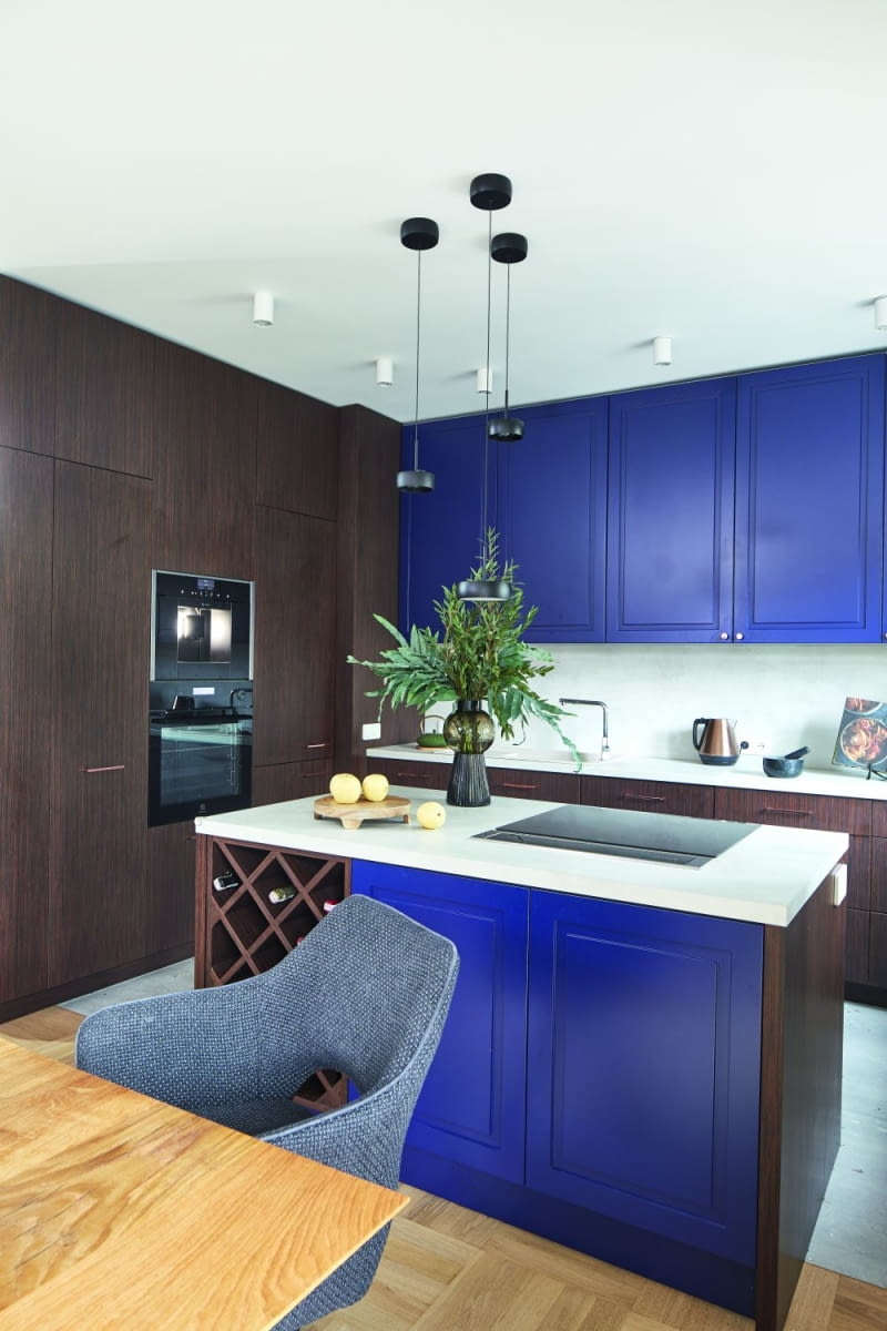 Jasną i przestronną strefę dzienną urozmaicają intensywne kolory mebli kuchennych. Ściana ciemnego, drewnianego forniru skrywa zabudowane sprzęty AGD, półki, ale także szacht kominowy w narożniku. Zdobi również część dolnych szafek i kontrastuje z niebieskimi frontami, dobranymi w tym samym odcieniu co ściana w salonie. Mocne kolory przełamują jasne, lekko szare barwy blatów oraz ściany między dolną i górną linią szafek wykończonych laminatem marki Egger. Na podłodze ułożono również jasne płytki podłogowe. Pochodzą od firmy Marmara i imitują lastryko z kolorowymi wtrąceniami.