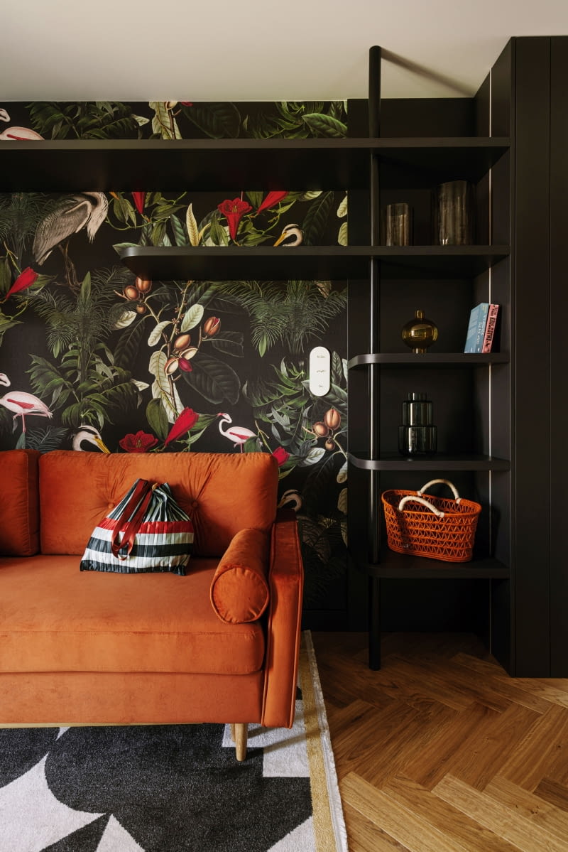 W gabinecie pojawiła się ruda sofa z miękkiego weluru – idealne miejsce na chwilę odpoczynku w trakcie pracy. Jej wyszukany charakter podkreśla ciemna tapeta z motywem roślinnym  i zwierzęcym ze sklepu Wallcolors. Ciepły odcień tapicerki powtórzono w dodatkach, m.in.  w pomarańczowym koszu z salonu NAP.