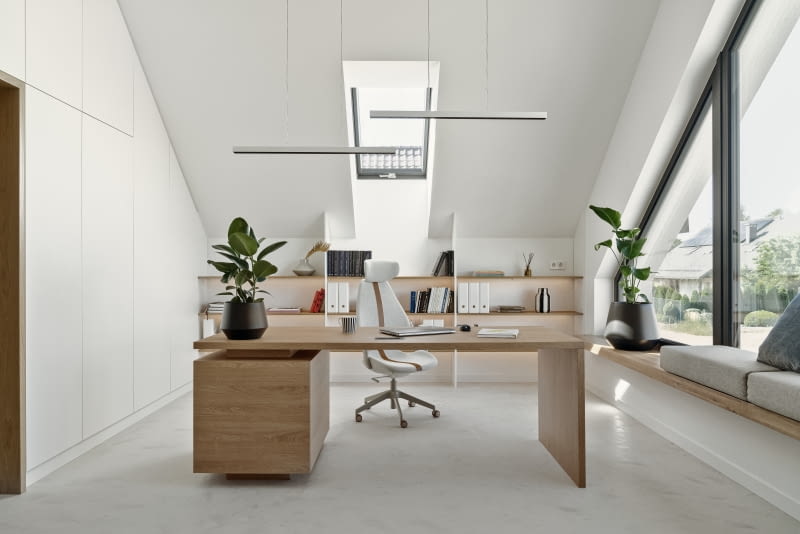 W gabinecie podłogę wykończono tak jak na parterze jasnoszarą żywicą. Pod jej powierzchnią w całym domu zainstalowano ogrzewanie podłogowe. Dzięki temu nie ma konieczności montażu pionowych grzejników, co byłoby kłopotliwe w pomieszczeniu z tak nisko osadzonym oknem jak gabinet. Na tle jasnej podłogi ładnie prezentują się drewniane elementy wykonane na zamówienie z płyty laminowanej w kolorze dębu – biurko i półki. Nad biurkiem zawisły te same lampy co nad stołem jadalnianym – polskiego producenta AQForm.